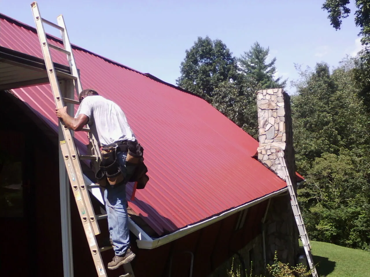 Roofer on a ladder beside a red metal roof and stone chimney — Soffit & Fascia Repair by Roofing Brothers in Watauga
